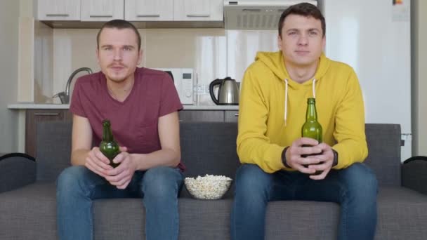 ソファに座っている2人の男性がテレビでサッカーの試合を見たり ビールを飲んだり ポップコーンを食べたりします 二人の若者が冷静にスポーツの試合を見ていた ポップコーンが床に落ちる背景にキッチン 4Kフッテージ — ストック動画