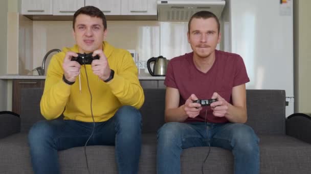 两个男人正在互相玩电子游戏 年轻人坐在坐在几把架上 手里拿着游戏垫 玩着游戏机 其中一人赢了 享受着胜利 第二个朋友很不高兴 — 图库视频影像