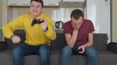 İki adam video oyunu oynuyor ve kazanıyor. Genç çocuklar kanepede oturuyorlar, ellerinde gamepad'ler tutuyorlar, konsol oynuyorlar, zaferin tadını çıkarıyorlar, bunlardan biri kanepeden kalkıyor ve kazanan bir dans yapıyor.