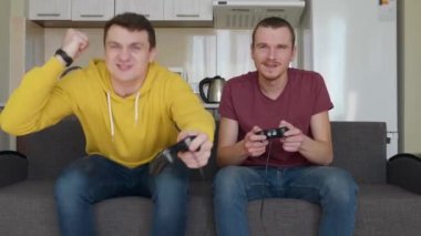 İki adam video oyunu oynar ve kaybeder. Genç çocuklar kanepede oturuyorlar, ellerinde gamepad'ler tutuyorlar ve erken kazandıklarını düşünüyorlar, ama sonunda kaybettiler. Arkadaşlar dinlensin. Arka plandaki mutfak. 4k görüntü.