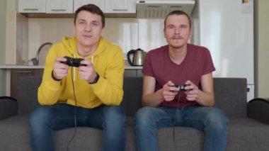 İki adam video oyunu oynuyor ve kazanıyor. Genç çocuklar kanepede oturuyor, ellerinde gamepads tutun, konsol oynamak, zafer zevk, bir arkadaş kanepeden yükselir ve ellerini sallar. Takım maçı. 4k görüntü.