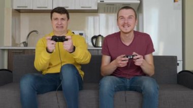 İki adam birbirlerine karşı bir video oyunu oynuyorlar. Genç çocuklar kanepede oturuyorlar, ellerinde gamepad'ler tutuyorlar ve, biri kazanıyor, kanepeden kalkıp seviniyor, ikinci arkadaş üzgün. 4k görüntü.