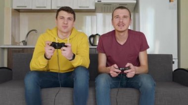 İki adam gülümsüyor ve coşkuyla bir video oyunu oynuyorlar. Genç mutlu çocuklar kanepede oturuyor, ellerinde gamepads tutun, konsol oynamak ve gülmek. Takım maçı. Arka planda mutfak. 4k görüntü.