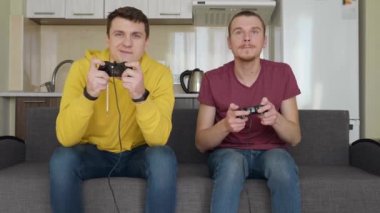 İki adam video oyunu oynuyor ve kaybediyor. Genç çocuklar kanepede oturuyor, ellerinde gamepads tutun ve konsol oynamak, düzeyleri nin geçişi sırasında hatalarını tartışmak. Takım maçı. 4k görüntü.