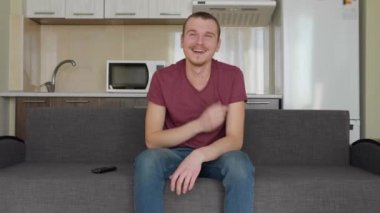 Bir adam televizyon izliyor. Kanepede oturan neşeli bir genç adam ve sert gülüyor. Arka planda bir mutfak. 4k görüntü.