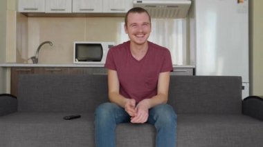 Bir adam televizyon izliyor. Kanepede oturan neşeli bir genç adam ve sert gülüyor. Arka planda bir mutfak. 4k görüntü.