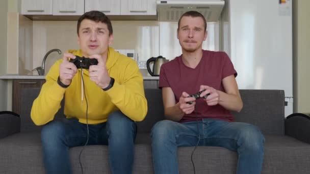二人の男がお互いにビデオゲームをしている 若い男はソファに座って 自分の手でゲームパッドを保持し コンソールを再生し そのうちの1つが勝つと勝利を楽しむ 第二の友人は4K映像を動揺しています — ストック動画
