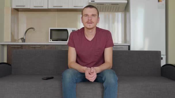 一个男人正在看电视 一个开朗的年轻人坐在沙发上 笑得很厉害 背景上的厨房 — 图库视频影像