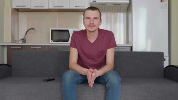一个男人正在看电视 年轻人坐在沙发上 用手捂着脸 背景上的厨房 — 图库视频影像