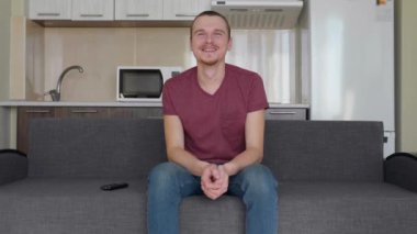 Bir adam televizyon izliyor. Kanepede oturan neşeli bir genç adam, gülüyor, televizyon seti parmağını işaret eder ve ellerini kötü alkışlar. Arka planda bir mutfak. 4k görüntü.