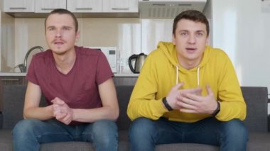 Kanepede oturan iki adam tv'de bir futbol maçı izlemek ve en sevdikleri takımın zaferinin tadını çıkarın. Adamlardan biri kanepeden kalkıp kollarını sallıyor. Arka planda mutfak var. 4k görüntü.