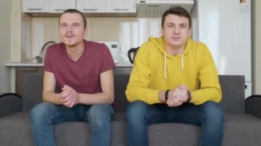 Kanepede oturan iki adam tv'de bir futbol maçı izlemek ve en sevdikleri takımın zaferinin tadını çıkarın. Genç çocuklar kanepeden kalkıp sevinçten birbirlerine sarılırlar. Arka planda mutfak var. 4k görüntü.