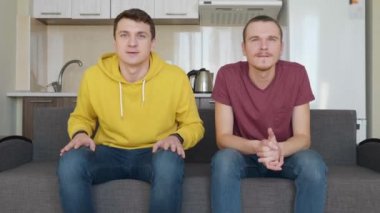 Kanepede oturan iki adam tv'de bir futbol maçı izlemek ve en sevdikleri takımın zaferinin tadını çıkarın. Genç çocuklar kanepeden kalkıp sevinçten birbirlerine sarılırlar. Arka planda mutfak var. 4k görüntü.