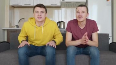 Kanepede oturan iki adam televizyonda bir futbol maçı izlemek ve farklı takımlara destek. Sevincini kazanan takımı destekleyen adam ve kaybeden takımın hayranı olan arkadaşı, elleriyle yüzünü kapatıyor. 4k görüntü.