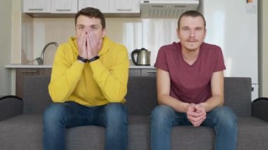 Kanepede oturan iki adam televizyonda bir futbol maçı izlemek ve farklı takımlara destek. Sevincini kazanan takımı destekleyen adam ve kaybeden takımın hayranı olan arkadaşı, elleriyle yüzünü kapatıyor. 4k görüntü.