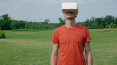 Parkta VR kulaklık kullanan bir adam ve kafasını açar. Kırmızı bir tişört ve bir kask genç bir adam doğanın arka planına karşı sanal gerçeklik yaşıyor. Doğru çerçevede bir insan. 4k görüntüleri.