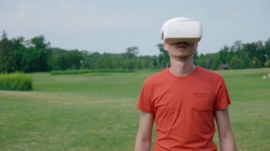 Parkta VR kulaklık kullanan bir adam ve kafasını açar. Kırmızı bir tişört ve bir kask genç bir adam doğanın arka planına karşı sanal gerçeklik yaşıyor. Doğru çerçevede bir insan. 4k görüntüleri.
