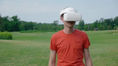 Parkta VR kulaklık kullanan bir adam ve kafasını açar. Kırmızı bir tişört ve bir kask genç bir adam doğanın arka planına karşı sanal gerçeklik yaşıyor. Doğru çerçevede bir insan. 4k görüntüleri.