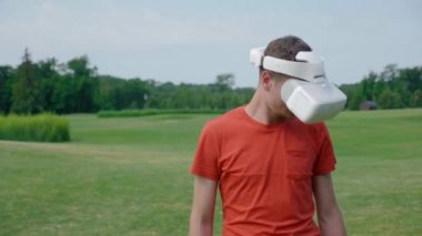 Parkta VR kulaklığı kullanan bir adam ve kafasını aşağı çevirir. Kırmızı bir tişört ve bir kask genç bir adam doğanın arka planına karşı sanal gerçeklik yaşıyor. Doğru çerçevede bir insan. 4k görüntüleri.