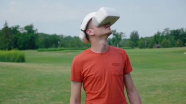 Parkta VR kulaklık kullanan bir adam ve kafasını açar. Kırmızı bir tişört ve bir kask genç bir adam doğanın arka planına karşı sanal gerçeklik yaşıyor. Doğru çerçevede bir insan. 4k görüntüleri.