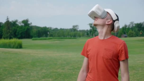 公園でVrヘッドセットを使用して 彼の頭を上げる男 赤いTシャツとヘルメットを着た若い男が 自然を背景にバーチャルリアリティを体験しています フレームの右側の人間4Kフッテージ — ストック動画