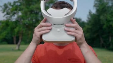 Bir adam bir park içinde kafasına VR kulaklık koyar. Kırmızı bir tişört ve bir kask genç bir adam doğanın arka planına karşı sanal gerçeklik yaşıyor. Çerçevenin merkezinde bir insan. Close. 4k görüntüleri.