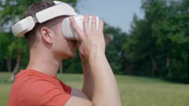 Bir adam onun VR kulaklığı parkta kafasını alır. Kırmızı bir tişört ve bir kask genç bir adam doğanın arka planına karşı sanal gerçeklik yaşıyor. Close. 4k görüntüleri.