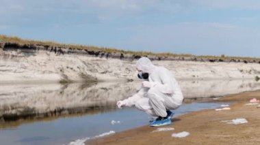 Koruyucu kostümlü ve solunum cihazı maskesi giymiş adam kirli nehre, göle gelir ve test tüpünde analiz için su alır. Bilim adamı örnek lerini in vitro olarak alır. Ekolojik felaket. 4k