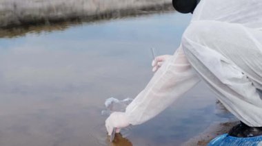 Koruyucu kostümlü ve solunum cihazı maskesi giymiş adam kirli nehre, göle gelir ve test tüpünde analiz için su alır. Bilim adamı örnek lerini in vitro olarak alır. Ekolojik felaket. 4k