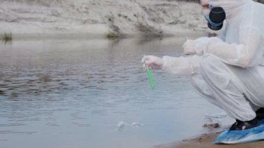 Koruyucu kostümlü ve solunum cihazına ait gaz maskesi giymiş adam, kirli bir nehirden, gölden analiz için aldığı test tüpündeki suyun kimyasal bir reaksiyonu gözlemliyor. Bilim adamı örnek lerini in vitro olarak alır. 4k