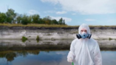 Koruyucu giysili ve solunum cihazı giyen bir adam kameraya gidiyor ve kirli nehir veya gölden gelen su analizleri içeren bir test tüpü gösteriyor. Bilim adamı örnek lerini in vitro olarak alır. Ekolojik felaket. 4k