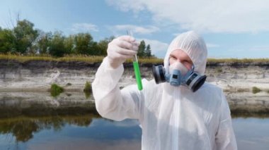 Koruyucu giysili ve solunum cihazı giyen adam test tüpündeki suyun kimyasal reaksiyonuna dikkat eder, ki bunu kirli nehirden analiz etmek için aldı, sonra da bırakır. Bilim adamı örnek lerini in vitro olarak alır. 4k görüntü.
