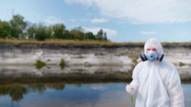 Koruyucu giysili ve solunum cihazı giyen bir adam kameraya gidiyor ve kirli nehir veya gölden gelen suyun analizini içeren bir test tüpü gösteriyor. Bilim adamı örnek lerini in vitro olarak alır. Ekolojik felaket. 4k