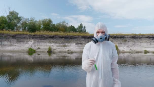 防護服を着た男と人工呼吸器のガスマスクは 汚染された川 湖の背景に対して彼の手に試験管を持って立って カメラを見ます 生態学的な大きなトロフィー4K映像 — ストック動画