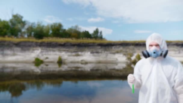 防護服を着た男と人工呼吸器がカメラに向かって 汚染された川や湖からの水の分析を含む試験管を示しています 科学者はインビトロでサンプルを採取します 生態学的災害4K — ストック動画