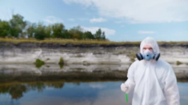 防護服を着た男と人工呼吸器がカメラに向かって 汚染された川や湖からの水の分析を含む試験管を示しています 科学者はインビトロでサンプルを採取します 生態学的災害4K — ストック動画