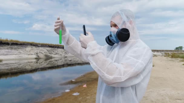 防護服を着た男性と人工呼吸器を装着した男性が 川や湖の背景に試験管を持って立ち スマートフォンを使って水の化学反応を観察している 4Kフッテージ — ストック動画