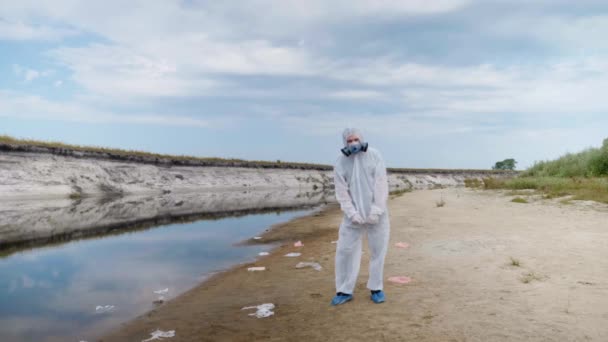 一个穿着防护服和呼吸器的男子站在塑料垃圾中间 靠近一条干涸的河流 挥舞着双手 人类正在毒害地球 生态灾难 人为影响 — 图库视频影像