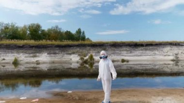 Koruyucu giysili ve solunum cihazı giyen bir adam, kurumuş bir nehrin yanında plastik çöpler arasında duruyor ve ellerini sallıyor. İnsanlık Dünya gezegenini zehirliyor. Ekolojik felaket. Antropojenik etki. 4k