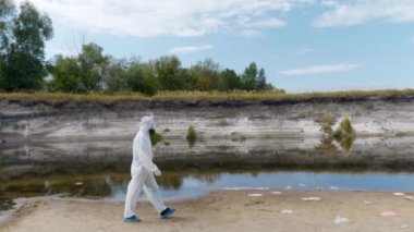 Koruyucu giysili ve solunum cihazı giyen bir adam kuru bir nehir boyunca yürür ve kirliliğin boyutunu tahmin ediyor. İnsanlık Dünya gezegenini zehirliyor. Ekolojik felaket. Antropojenik etki. 4k