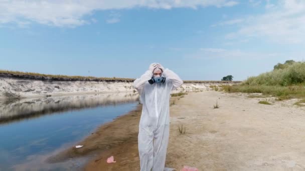 防護服を着た男と人工呼吸器が乾いた川に沿って歩き 頭を手に取り 汚染の程度を推定する 生態学的災害人為的影響4Kフッテージ — ストック動画