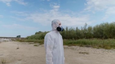 Koruyucu giysili ve solunum cihazı giyen bir adam, kurumuş bir nehrin yakınında plastik çöplerin arasında duruyor ve kirliliğin boyutunu tahmin ediyor. İnsanlık Dünya gezegenini zehirliyor. Ekolojik felaket. 4k