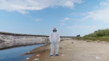 Koruyucu giysili adam ve solunum cihazı nehrin plastik kirli kıyıları arasında duruyor, göl başını kaldırır ve kameraya bakar. Ekolojik felaket. Antropojenik etki. Gezegeni kurtar. 4k