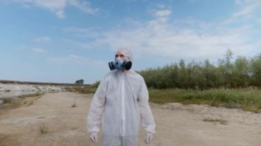 Koruyucu giysili ve solunum cihazı giyen adam, insanların plastik çöp bıraktığı ve kirliliğin boyutunu tahmin ettiği kurumuş bir nehrin yanındaki sahilde duruyor. Ekolojik felaket. Gezegeni kurtar. 4k