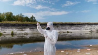 Koruyucu giysili ve solunum cihazı giyen adam, kirli nehirden analiz için aldığı test tüpündeki suyun kimyasal reaksiyonuna dikkat çekti. Bilim adamı örnek lerini in vitro olarak alır. Ekolojik felaket. 4k