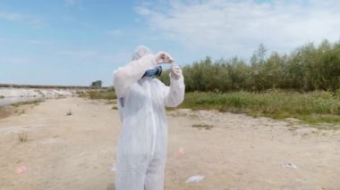 Koruyucu giysili ve solunum cihazı giyen adam, kirli nehirden analiz için aldığı test tüpündeki suyun kimyasal reaksiyonuna dikkat çekti. Bilim adamı örnek lerini in vitro olarak alır. Ekolojik felaket. 4k