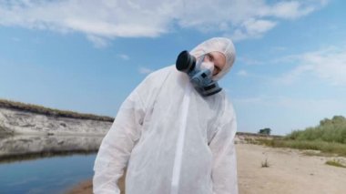 Koruyucu giysili ve solunum cihazı giyen adam, nehrin, gölün plastik kirli kıyıları arasında duruyor. Çerçeve döner. Ekolojik felaket. Antropojenik etki. Gezegeni kurtar. 4k görüntü.