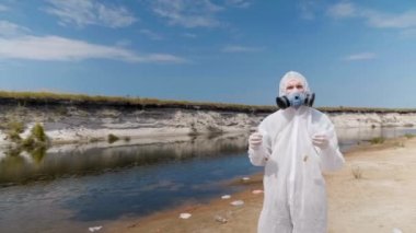 Koruyucu giysili ve vitro lu solunum cihazı giyen adam, insanların plastik çöp leri bırakıp kameraya baktığı kurumuş nehrin yanındaki plajda duruyor. Ekolojik felaket. Antropojenik etki. 4k