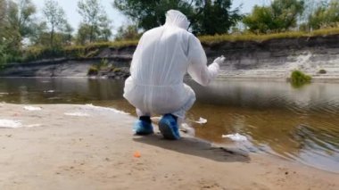Koruyucu giysili adam ve solunum cihazı kirli nehir, göl tarafından çömelmiş ve test tüpünde analiz için su alır. Bilim adamı örnek lerini in vitro olarak alır. Ekolojik felaket. Antropojenik etki. 4k