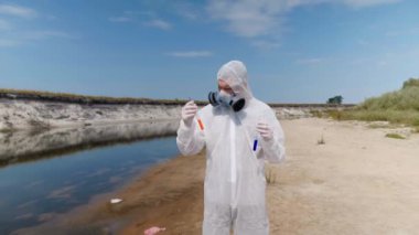 Koruyucu giysili ve solunum cihazı giyen adam, kirli nehirden analiz için aldığı test tüpündeki suyun kimyasal reaksiyonuna dikkat çekti. Bilim adamı örnek lerini in vitro olarak alır. Ekolojik felaket. 4k
